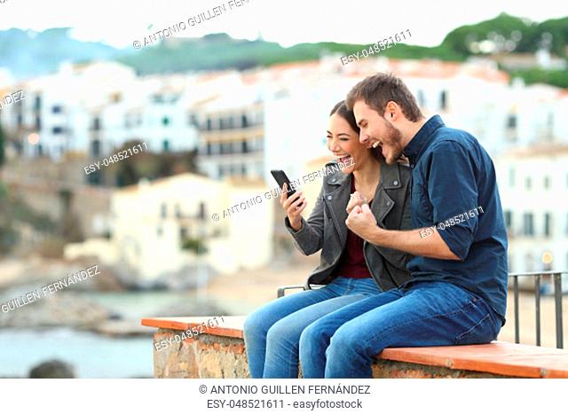 Excited couple finding news on smart phone on a ledge with a town in the background