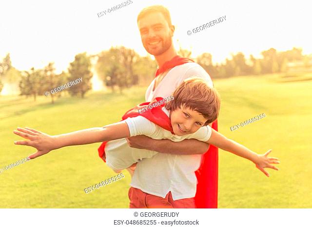 Handsome young dad and his cute little son dressed like superheroes are looking camera and smiling while playing in park