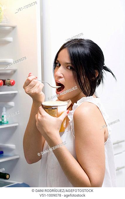 Brunette woman enjoying ice cream in the kitchen beside the fridge
