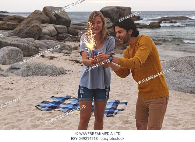 Front view of young Caucasian couple playing with fire cracker while standing at beach. They are smiling
