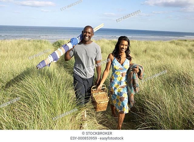 Young Couple Carrying Picnic Basket And Windbreak Walking Through Dunes