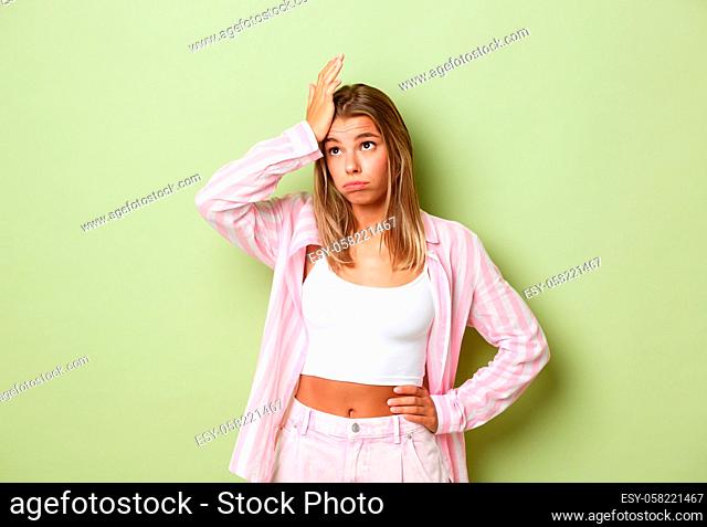 Portrait of exhausted and troubled blond business woman in stylish outfit, exhaling tired and holding hand on forehead, standing over green background
