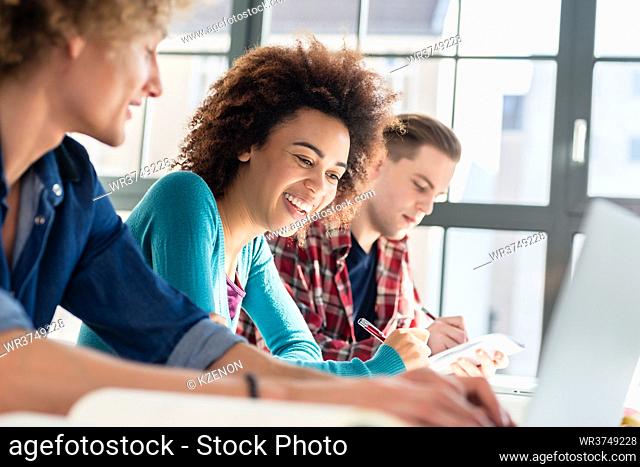Cheerful young woman writing an assignment while sitting at desk between two classmates during class at college or university