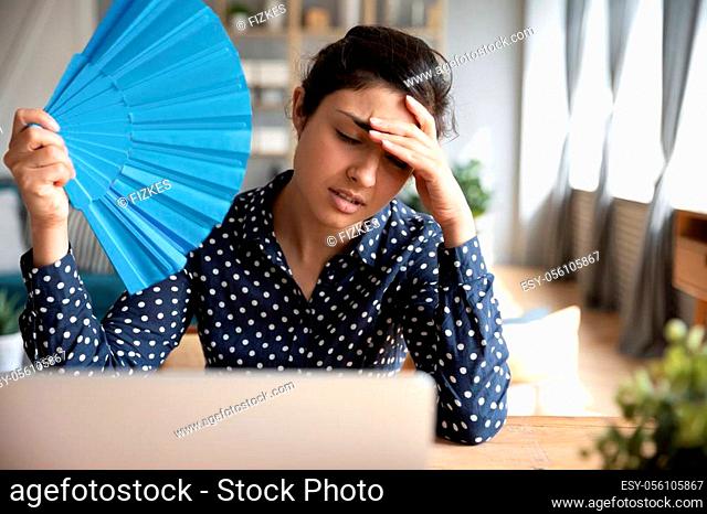 Tired overheated young indian woman hold wave fan suffer from heat sweating indoor work on laptop at home office, annoyed girl feel uncomfortable hot summer...