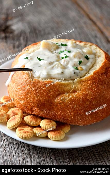 Vertical closeup of a bread bowl of New England Clam Chowder on a rustic wood table with a spoon and oyster crackers