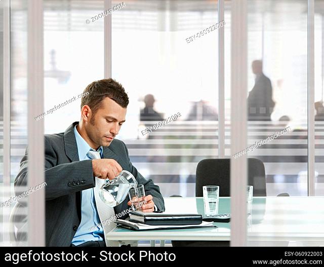 Businessman waiting in meeting room in modern, corporate office. Pouring water to glass