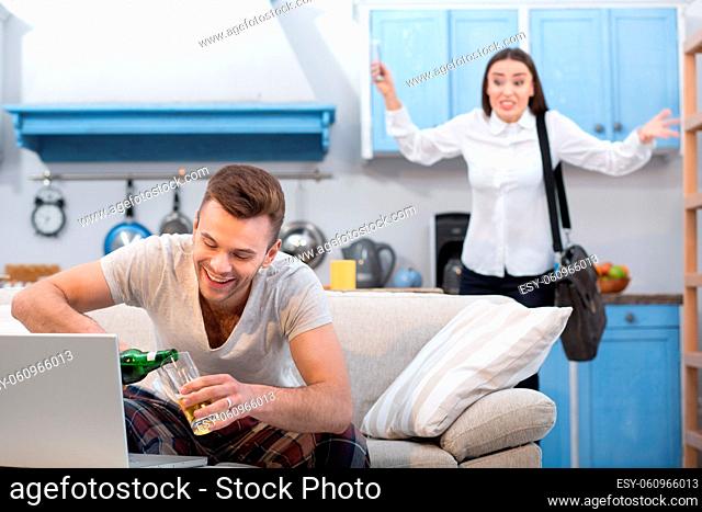 Woman going to work fights her unreliable husband. Man working at home, enjoying his time and drinking beer at couch