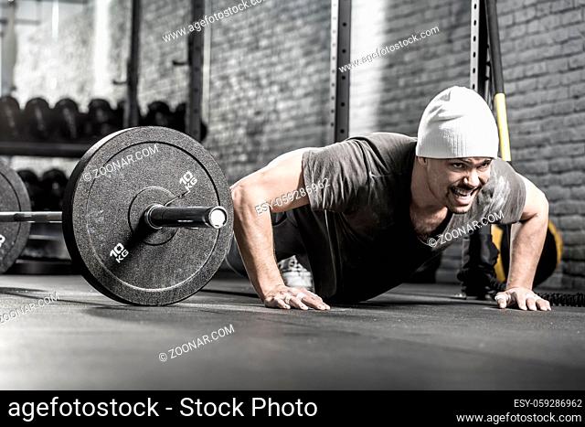 Muscular guy with a beard makes a pushup in the gym on the gray brick wall background. He wears sportswear, white sneakers and a white cap