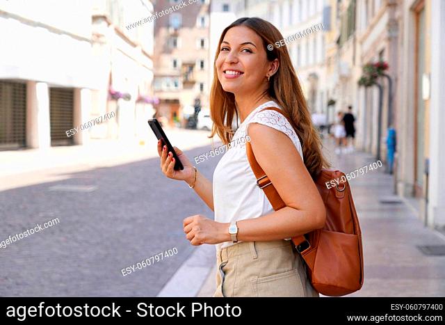 Pretty young woman walking in city street while holding the smartphone and looking sideways