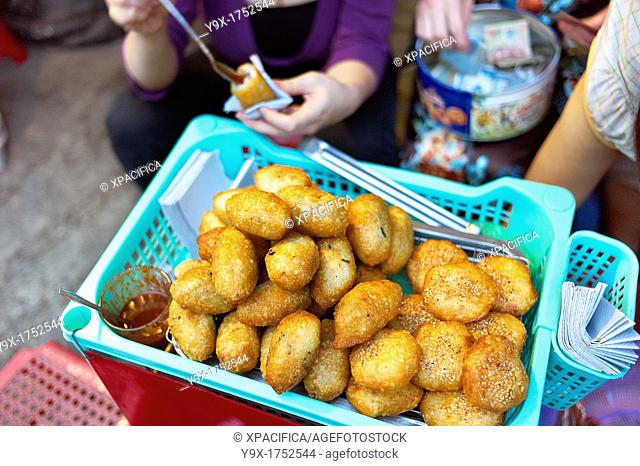 A woman selling the fried food banh ran, or deep fried rice ball, on the side of a street on makeshift displays and stools