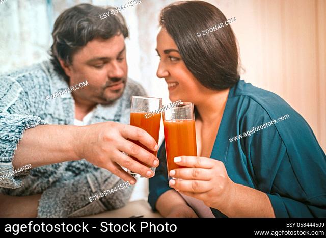 Body Positive Man And Woman Clinking With Glasses Of Orange Juice, Caucasian Couple Casual Clothing Flirting And Joining With Glasses While Sitting At Home