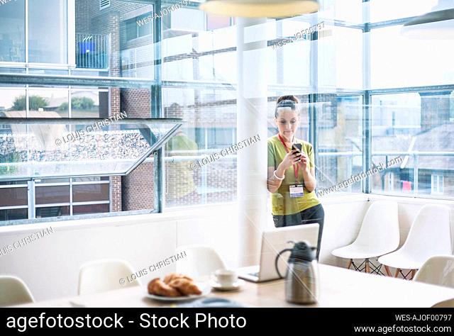 Young businesswoman text messaging on mobile phone while leaning on column at office