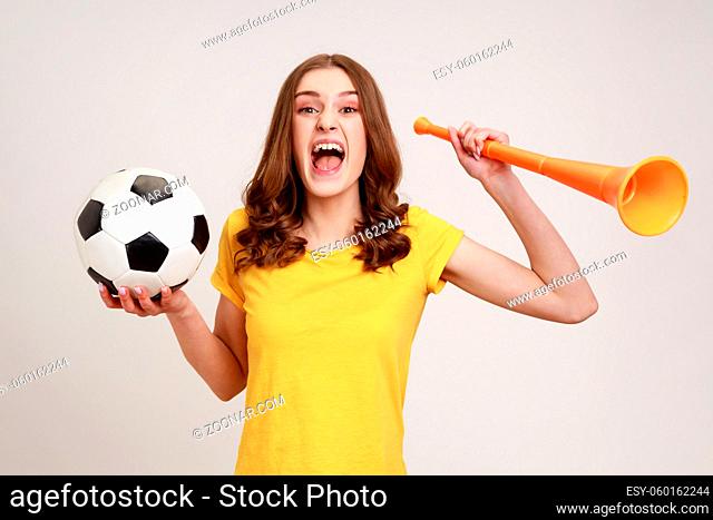 Portrait of cheerful woman of young age with brown hair in yellow t-shirt holding big bullhorn and football ball, celebrating championship beginning