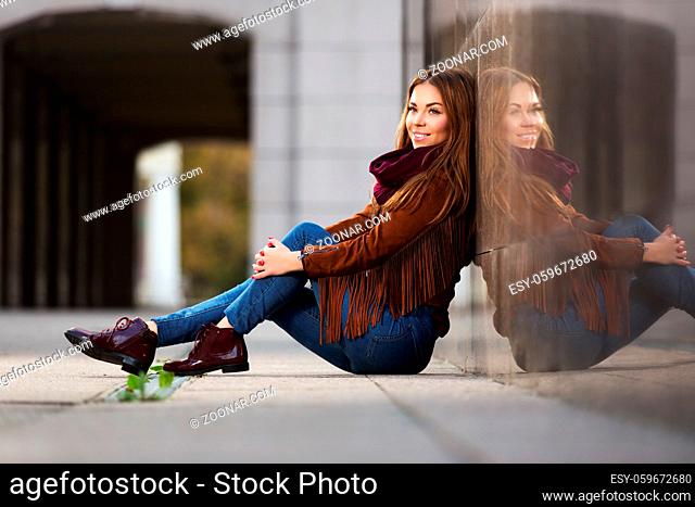 Happy young fashion woman in leather jacket sitting on city street