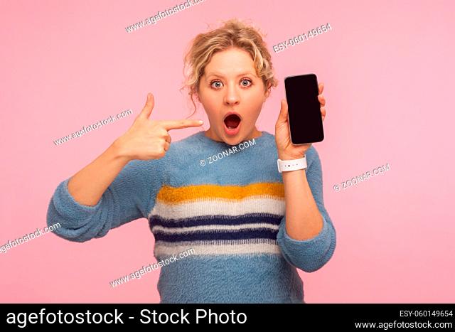 Wow, amazing smartphone. Astonished woman in warm sweater pointing to mobile phone and staring at camera with amazed face, shocked by cell phone or application