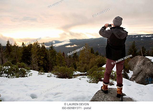 Mid adult man photographing view from rock on snow covered mountain, rear view, Twain Harte, California, USA