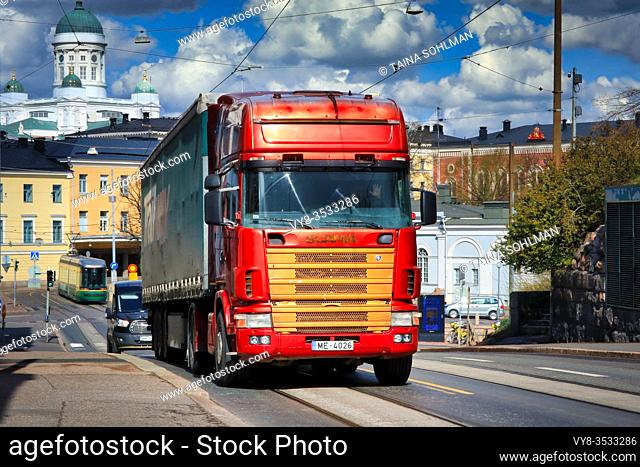 Red and gold 4-series Scania truck pulling semi trailer to Katajanokka Harbour. On the background city of Helsinki, Finland. April 29, 2020