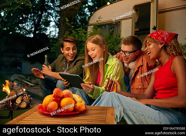 Happy friends spending evening nearby bonfire and campsite. Four millennial people watching funny video using mobile tablet smiling and laughing