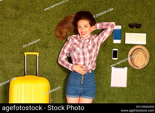 Portrait of toothy smiling beautiful tourist lady lying on green grass near yellow luggage, mobile or smart phone, passport, cap, map or guide, etc