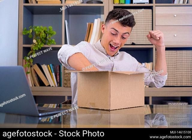 Smiling man wearing glasses unpacking awaited parcel, looking inside, sitting at work desk, satisfied happy customer opening cardboard box with online store...