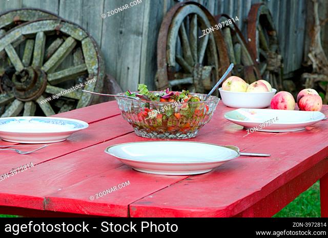 Glass dish full of salad and ecologic apples on wooden table in rural homestead. Wooden cart wheels on background