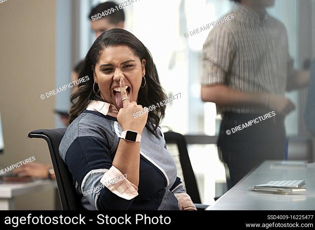 Young woman sitting at computer in office looking to the camera making funny face, co-workers having discussion around dry erase board in background