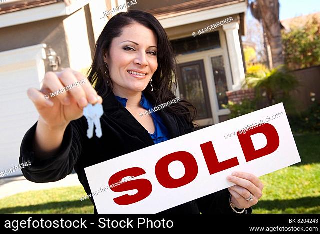 Happy attractive hispanic woman holding keys and sold sign in front of house