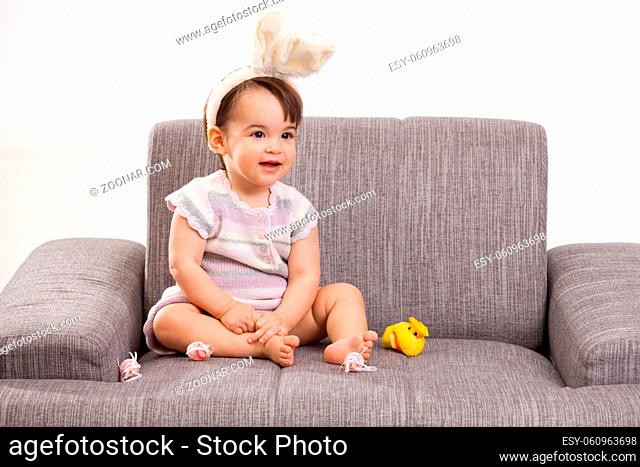 Baby girl in easter bunny costume, sitting on grey couch playing with toy chicken and easter eggs, laughing. Isolated on white background