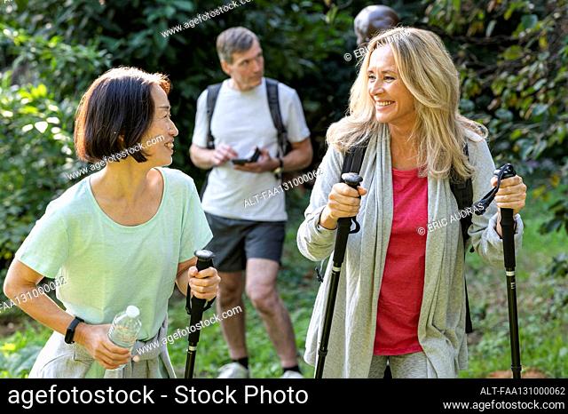 Two middle-aged ladies with hiking poles walking in the woods with their partners behind them