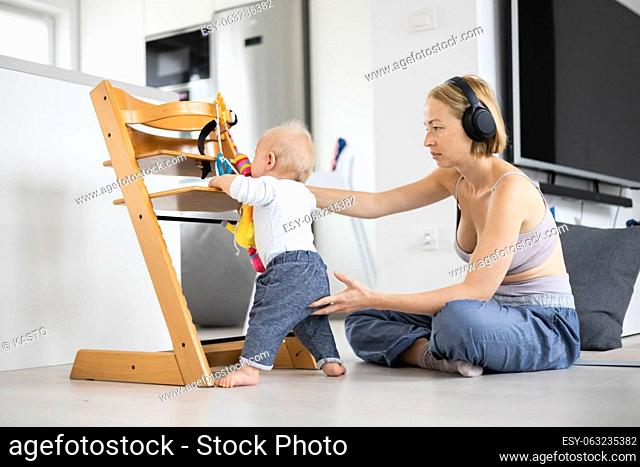 Women's multitasking. Mother sitting on floor playing with her baby boy watching and suppervising his first steps while listening to podcast on wireless...