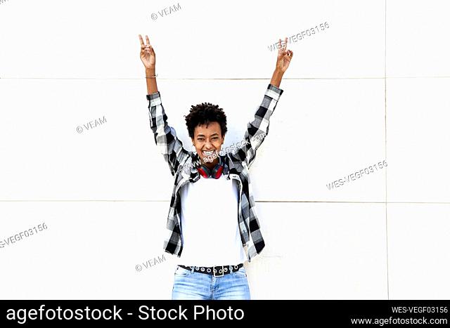 Cheerful female Afro hipster showing peace sign with arms raised against white wall