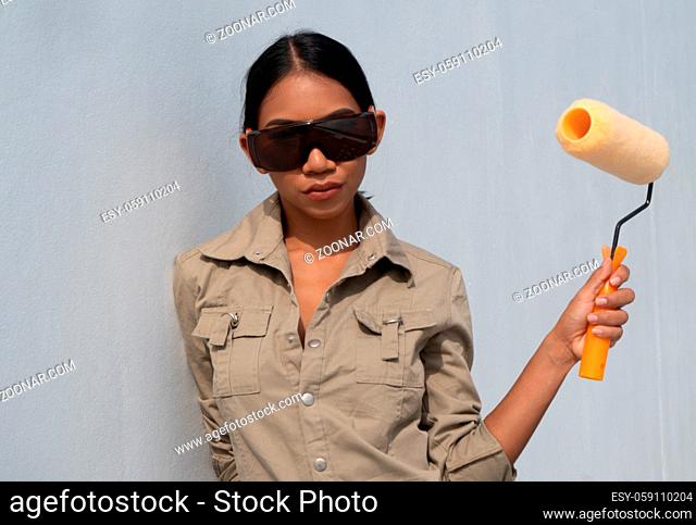 Portrait of attractive young girl builder with paint roller in overall and protective glasses standing over white wall background