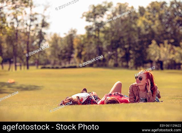 Portrait of happy best friends resting and spending free time in park. Beautiful lady in sunglasses looking at camera while her friends lying on rug