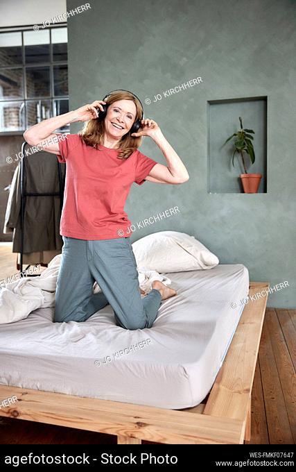Smiling woman wearing wireless headphones kneeling on bed at home