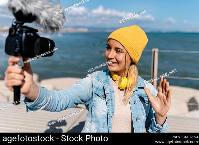 Smiling woman in knit hat waving hand while vlogging through camera during weekend