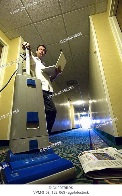 Low angle view of a businessman holding a laptop and a vacuum cleaner in a corridor