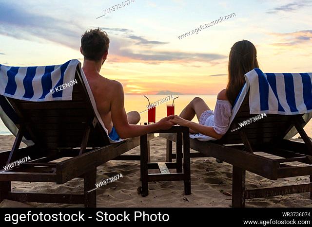 Rear view of a young romantic couple in love sitting on wooden chairs, while drinking cocktails on a tropical beach at sunset during vacation or honeymoon in...