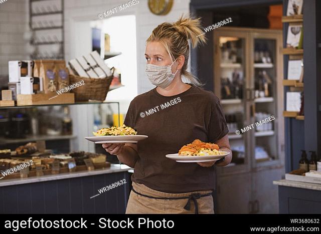 Blond waitress wearing face mask working in a cafe, carrying plates of food