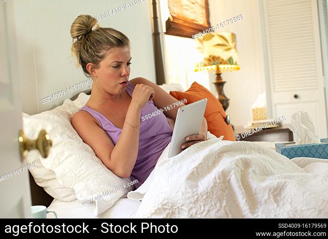 Coughing young woman in her bed with box of tissue using tablet to check in with doctor