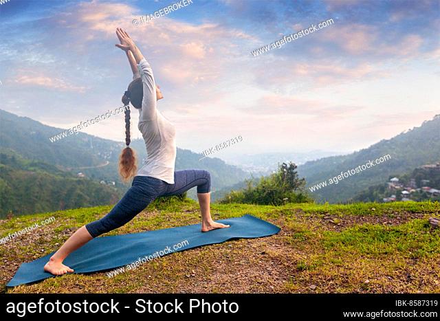 Yoga outdoors, sporty fit woman doing Ashtanga Vinyasa Yoga asana Virabhadrasana 1 Warrior pose posture in HImalayas mountains on sunset