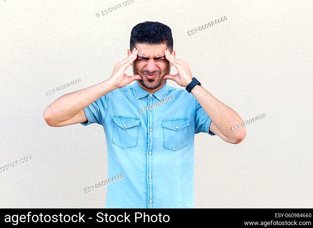 headache, confusion or problem. Portrait of sick handsome young bearded man in blue shirt standing and holding his head and feeling bad