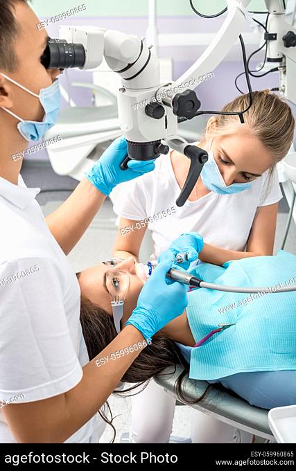 Patient in protective glasses on chair and dentist with assistant who sit next to her. Man looks on her teeth using a dental microscope and holds dental...
