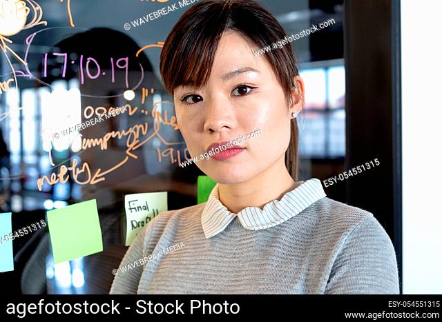 Portrait of an Asian female business creative working in a casual modern office, standing in front a glass wall with notes on it, looking straight to camera