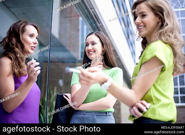 Female colleagues gossiping while smoking cigarettes during break outside office