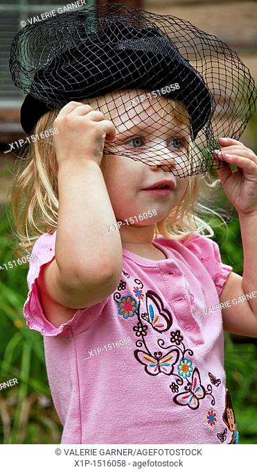 This vertical image shows a cute 2 year old Caucasian girl, wearing a black vintage hat with a veil over her face