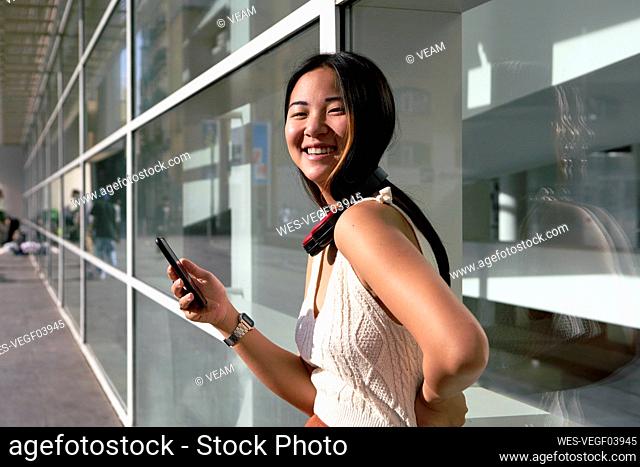 Smiling woman with smart phone and headphones around neck against glass wall