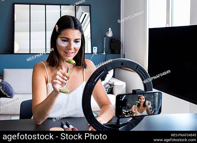 Young woman with eye mask showing jade stone roller on mobile phone at home