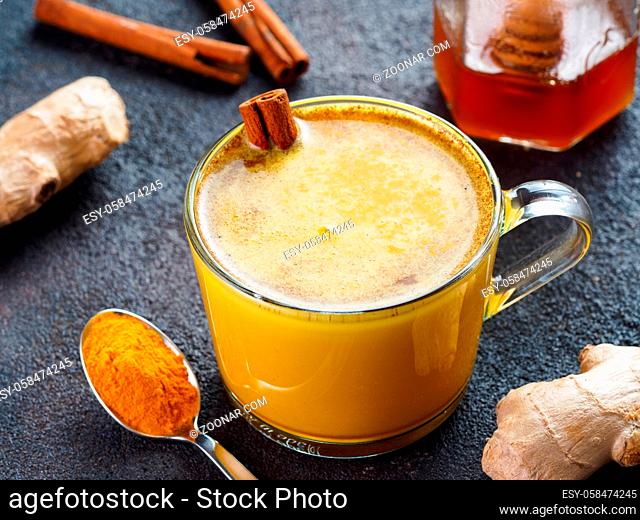 Healthy drink golden turmeric latte in glass cup.Gold milk with turmeric, ginger root, cinnamon sticks, dried turmeric powder and honey over black cement...
