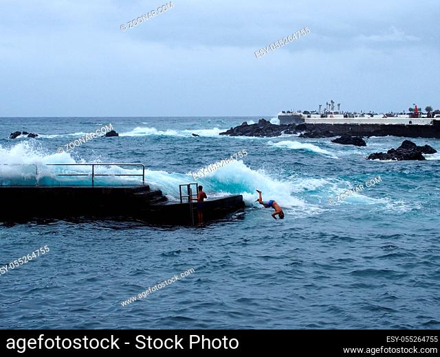 young man jumping off a concrete pier into the sea with high waves and white surf in puerto de la cruz tenerife