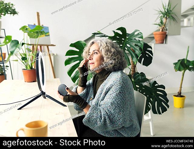 Senior woman applying foundation make-up in front of ring light at home